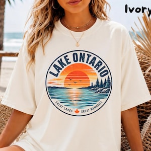 Peut inclure: Un t-shirt à manches courtes ivoire avec un motif circulaire représentant le lac Ontario. Le design inclut un coucher de soleil sur l'eau, des pins, des oiseaux et le texte 'LAKE ONTARIO GREAT LAKES GREAT MEMORIES' avec une petite feuille d'érable orange.