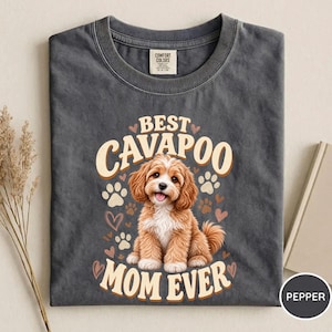 May include: A charcoal grey t-shirt featuring a graphic of a light brown Cavapoo puppy sitting in the centre. The text reads BEST CAVAPOO MOM EVER in a cream-coloured font, surrounded by small heart and paw print illustrations in cream and brown.