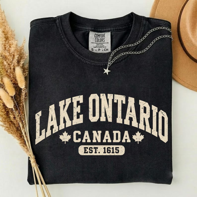 May include: A black crewneck t-shirt featuring the text LAKE ONTARIO CANADA EST. 1615 in a distressed cream font. The design includes two maple leaf icons and a Comfort Colors brand label at the inner neckline. A silver star necklace rests on top.