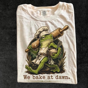 Peut inclure: Un t-shirt couleur crème avec une illustration représentant une grenouille verte portant une toque et un tablier de chef, tenant un rouleau à pâtisserie en bois et un bol avec un fouet. Le texte en bas indique, We bake at dawn. Étiquette Comfort Colors.