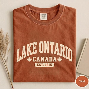 May include: A rust-coloured short-sleeve t-shirt with a crew neckline. The front features distressed white text that reads LAKE ONTARIO CANADA EST. 1615, flanked by two small maple leaf icons. The shirt has a Comfort Colors brand label at the neck.