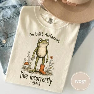 May include: An ivory t-shirt featuring a graphic of a green frog wearing one orange boot, standing next to a mushroom and wildflowers. The text reads, I'm built different like incorrectly i think. The shirt has a Comfort Colors brand label.