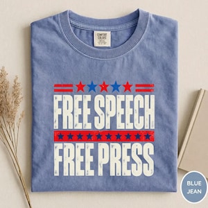 May include: A blue crew neck t-shirt with a graphic print featuring the text FREE SPEECH FREE PRESS in white capital letters. The design includes red and blue stars and horizontal lines, with a Comfort Colors brand label at the inner neckline.