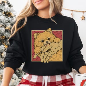 K&ouml;nnte beinhalten: Schwarzes Sweatshirt mit einer Cartoon-Illustration eines Zwergspitzes, der in eine Hand bei&szlig;t. Der Hund ist hellbraun, mit einem roten und gelben Hintergrund. L&auml;ssiger Stil.