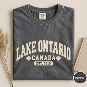 May include: A charcoal grey short-sleeve t-shirt featuring the text LAKE ONTARIO CANADA EST. 1615 in a distressed cream font. The design includes two small maple leaf icons flanking the word CANADA on the front of the garment.
