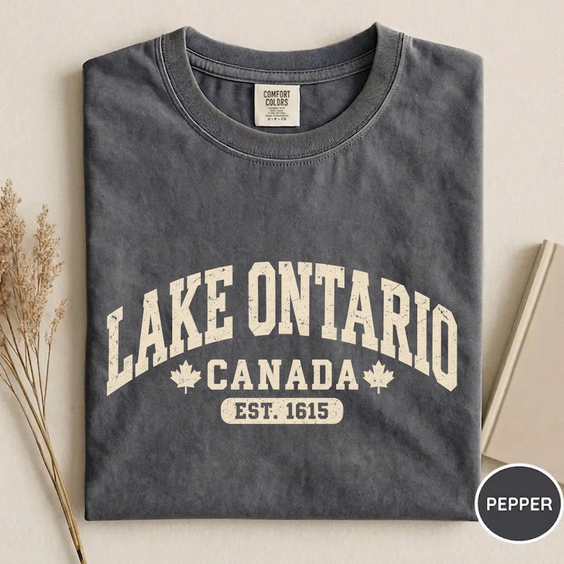 May include: A charcoal grey short-sleeve t-shirt featuring the text LAKE ONTARIO CANADA EST. 1615 in a distressed cream font. The design includes two small maple leaf icons flanking the word CANADA on the front of the garment.