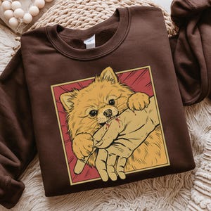 K&ouml;nnte beinhalten: Ein braunes Sweatshirt mit einer Grafik eines Zwergspitzes, der in eine Hand bei&szlig;t. Der Hund ist hellorange mit einem roten und gelben Hintergrund. Die Hand hat Blut. Das Sweatshirt hat eine einfarbige braune Farbe.