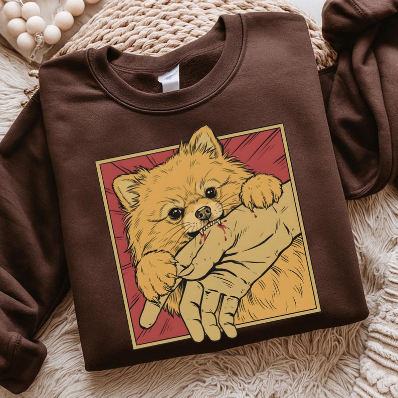 K&ouml;nnte beinhalten: Ein braunes Sweatshirt mit einer Grafik eines Zwergspitzes, der in eine Hand bei&szlig;t. Der Hund ist hellorange mit einem roten und gelben Hintergrund. Die Hand hat Blut. Das Sweatshirt hat eine einfarbige braune Farbe.