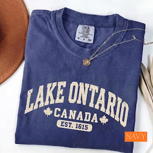 May include: A navy blue short-sleeved t-shirt with a crew neckline and a Comfort Colors brand label. The front features cream-coloured text reading LAKE ONTARIO CANADA EST. 1615 with two maple leaf icons. A thin gold-toned chain necklace rests on the shirt.