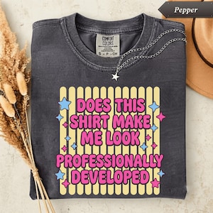 Peut inclure: Un t-shirt gris anthracite à manches courtes avec un col rond. Le motif central présente des rayures verticales jaunes avec le texte rose, Does This Shirt Make Me Look Professionally Developed, entouré de petites étoiles roses et bleues.