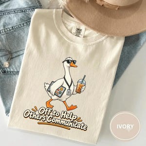 Puede incluir: Una camiseta de color marfil con un pato de dibujos animados que lleva gafas de sol y un cordón, sosteniendo una tableta y un café helado. El texto "Off to Help Others Communicate" está impreso debajo del pato en una fuente oscura y negrita.