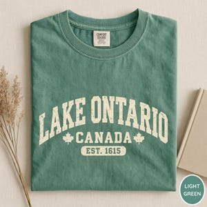 May include: A sage green crew neck t-shirt featuring the white text LAKE ONTARIO CANADA EST. 1615 with two maple leaf icons. The shirt has a Comfort Colors brand label at the inner neckline and a soft, washed-out fabric texture throughout the garment.