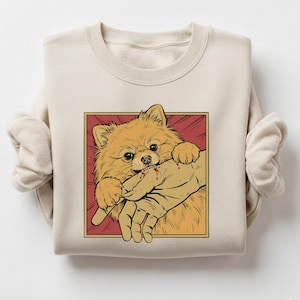 K&ouml;nnte beinhalten: Beiges Sweatshirt mit einer Cartoon-Illustration eines Zwergspitzes, der in eine Hand bei&szlig;t. Der Hund ist orange und braun, vor einem roten Hintergrund mit goldenem Rand. Die Hand hat etwas Blut.