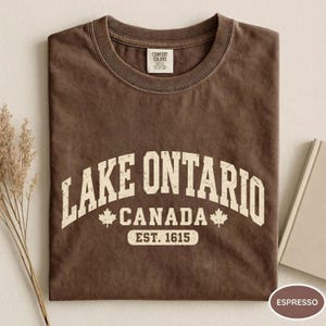 May include: A brown crew neck t-shirt featuring the white text LAKE ONTARIO CANADA EST. 1615 with two maple leaf icons. The shirt has a Comfort Colors brand label on the inside collar and a distressed, vintage-style graphic print on the front.