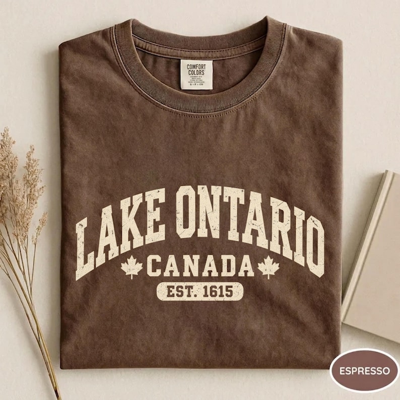 May include: A brown crew neck t-shirt featuring the white text LAKE ONTARIO CANADA EST. 1615 with two maple leaf icons. The shirt has a Comfort Colors brand label on the inside collar and a distressed, vintage-style graphic print on the front.