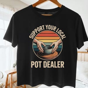Peut inclure: T-shirt noir avec l'inscription "SUPPORT YOUR LOCAL POT DEALER". Le graphisme représente des mains façonnant un pot sur un tour de potier, avec un coucher de soleil rétro en arrière-plan.