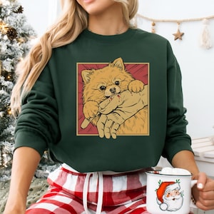 K&ouml;nnte beinhalten: Dunkelgr&uuml;nes Sweatshirt mit einer Cartoon-Illustration eines Zwergspitzes, der in eine Hand bei&szlig;t. Der Hund ist hellbraun mit einem roten und gelben Hintergrund. Das Sweatshirt wird von einer Person getragen.