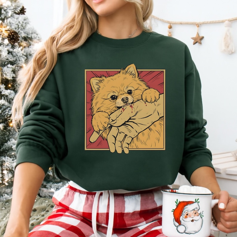 K&ouml;nnte beinhalten: Dunkelgr&uuml;nes Sweatshirt mit einer Cartoon-Illustration eines Zwergspitzes, der in eine Hand bei&szlig;t. Der Hund ist hellbraun mit einem roten und gelben Hintergrund. Das Sweatshirt wird von einer Person getragen.