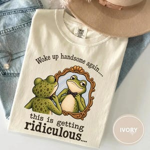 Peut inclure: Un t-shirt ivoire avec un motif représentant une grenouille verte portant un nœud papillon noir, se regardant dans un miroir doré orné. Le texte indique, Woke up handsome again... this is getting ridiculous.... Étiquette Comfort Colors.