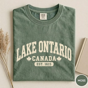 May include: A sage green crew neck t-shirt with a distressed white graphic print that reads LAKE ONTARIO CANADA EST. 1615, featuring two maple leaf icons. The shirt has a Comfort Colors brand label on the inside of the collar.