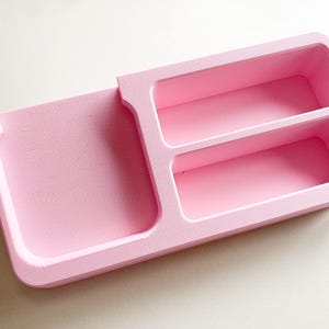 May include: A light pink organiser with a textured surface. It features a rectangular tray with two compartments and a separate, smaller tray. The organiser is designed to hold small items.