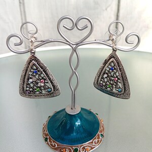 May include: Silver-toned triangle earrings with a textured border and colorful gemstones. The earrings are displayed on a decorative silver stand with a teal base and ornate detailing, showcasing a jewelry design.