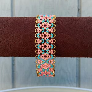 May include: A wide bracelet with a woven design. The bracelet features a pattern of pink and turquoise threads, connected by gold-colored metal links. The bracelet is displayed on a brown cylindrical surface.