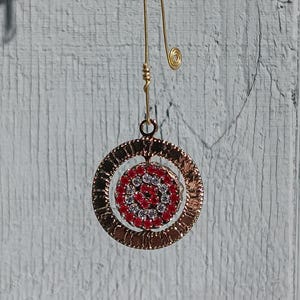 May include: Gold dangle earring with a round, textured, gold-toned metal design. The center of the design features a circle of red and white rhinestones.