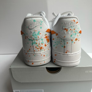 May include: Pair of white Nike Air Force 1 sneakers with teal and orange paint splatter design. The heels feature the Nike logo and "AIR" in white. The shoes are displayed on a gray shoe box.