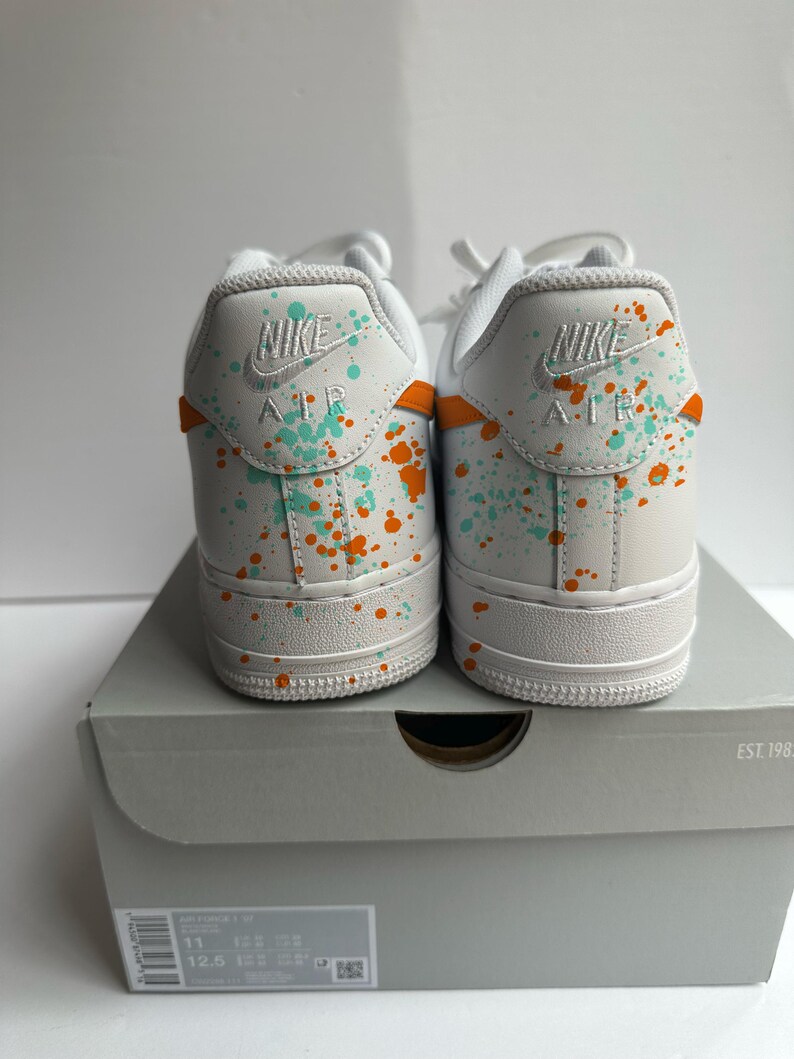 May include: Pair of white Nike Air Force 1 sneakers with teal and orange paint splatter design. The heels feature the Nike logo and "AIR" in white. The shoes are displayed on a gray shoe box.