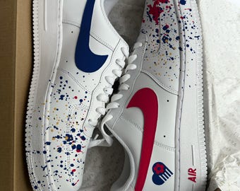 Puede incluir: Zapatillas blancas de cuero con logotipos Nike en azul y rojo. Las zapatillas tienen un fondo blanco con salpicaduras de pintura azul, roja y dorada. La zapatilla derecha tiene un corazón azul con rayas rojas y blancas en el lateral. La zapatilla izquierda tiene un corazón rojo con rayas azules y blancas en el lateral. La palabra "AIR" está impresa en la zapatilla derecha.