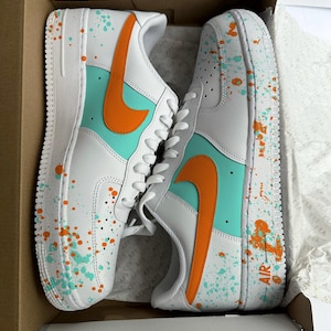 May include: White sneakers with orange and teal accents, featuring a splatter paint design. The shoes have an orange swoosh and teal panels. The word "AIR" is visible on the side of one shoe. The sneakers are in a cardboard box.