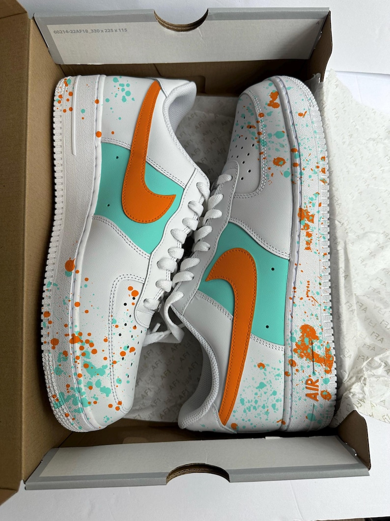 May include: White sneakers with orange and teal accents, featuring a splatter paint design. The shoes have an orange swoosh and teal panels. The word "AIR" is visible on the side of one shoe. The sneakers are in a cardboard box.