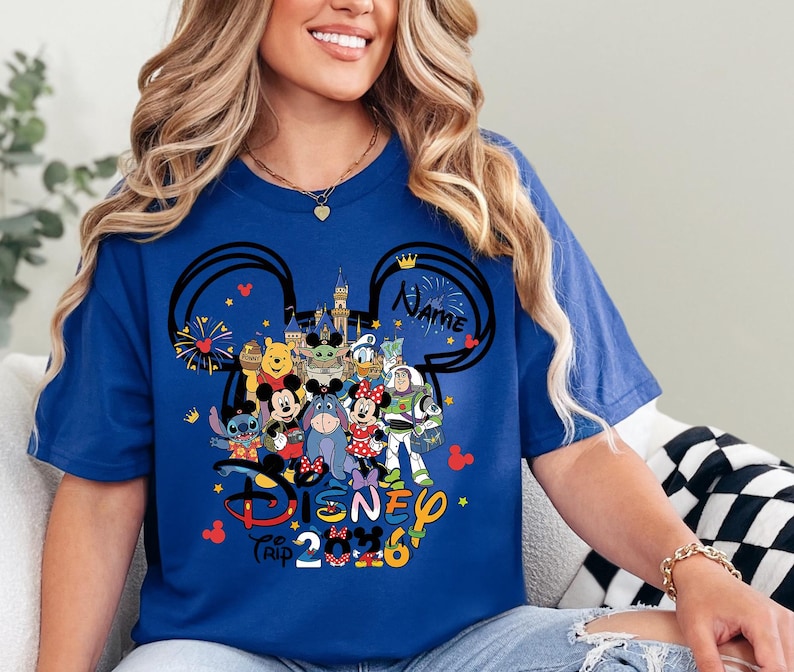 Peut inclure: T-shirt bleu roi avec un motif Disney Trip 2026. Le motif comprend des oreilles de Mickey Mouse, un ch&acirc;teau et divers personnages Disney tels que Mickey, Minnie et Winnie l'ourson. Le mot "Name" est &eacute;galement inclus.