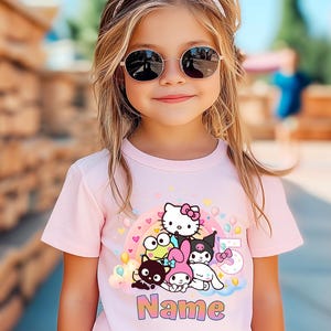 May include: A young girl wearing a pink t-shirt featuring a colorful graphic of cartoon characters, a rainbow, and the number 5. The shirt also has the word "Name" printed on it. She is wearing sunglasses and a pink bow.