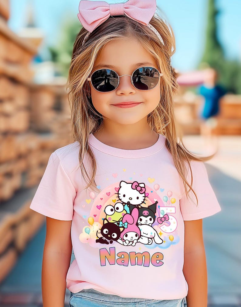 May include: A young girl wearing a pink t-shirt featuring a colorful graphic of cartoon characters, a rainbow, and the number 5. The shirt also has the word "Name" printed on it. She is wearing sunglasses and a pink bow.