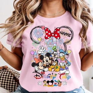 May include: Pink t-shirt featuring a Disney Trip design with Mickey Mouse, Minnie Mouse, Donald Duck, Goofy, and Pluto. The design includes a castle, fireworks, and the text "Disney Trip 2020". A red bow is at the top.