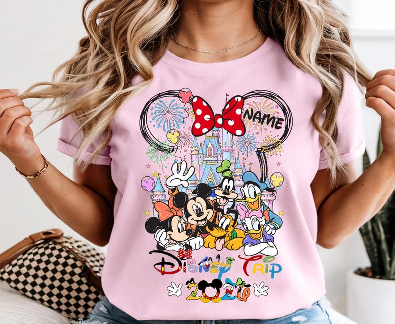May include: Pink t-shirt featuring a Disney Trip design with Mickey Mouse, Minnie Mouse, Donald Duck, Goofy, and Pluto. The design includes a castle, fireworks, and the text "Disney Trip 2020". A red bow is at the top.