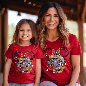Peut inclure: T-shirts rouges avec un motif inspir&eacute; de Minnie Mouse, repr&eacute;sentant divers personnages Disney et le texte "Disney 2023". Le motif comprend un n&oelig;ud &agrave; pois, des feux d'artifice et des personnages populaires.