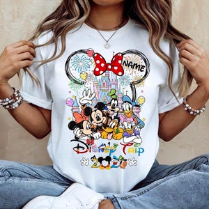 May include: White t-shirt featuring a colorful graphic of Disney characters, including Mickey and Minnie Mouse, Donald Duck, and Goofy. The design includes a castle, fireworks, and the words "Disney Trip 2020". A red bow with white polka dots is at the top.