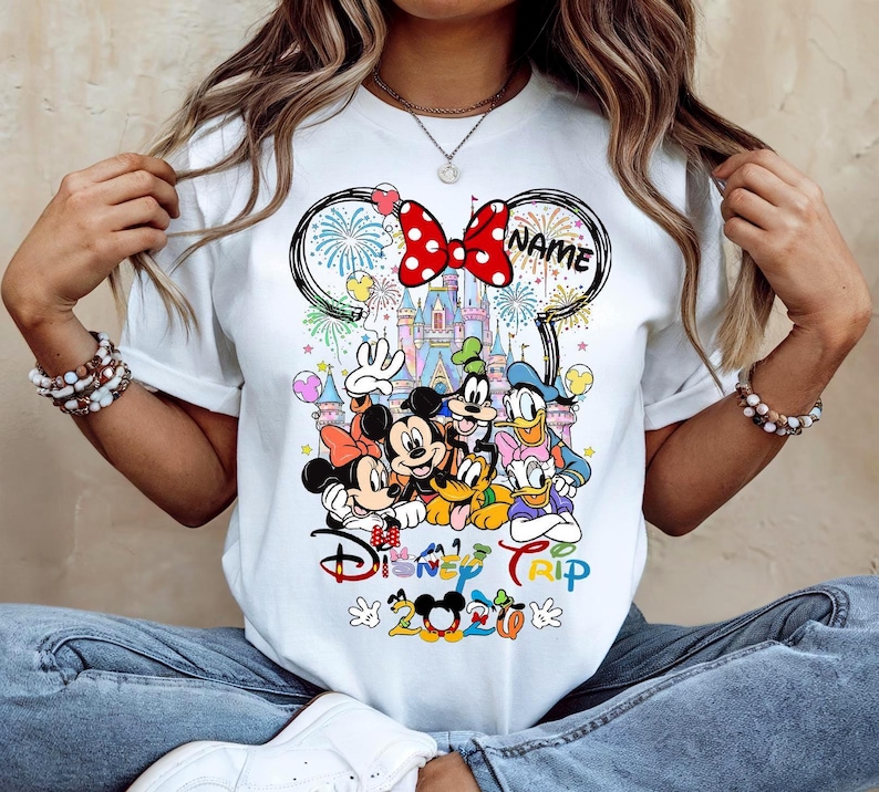 May include: White t-shirt featuring a colorful graphic of Disney characters, including Mickey and Minnie Mouse, Donald Duck, and Goofy. The design includes a castle, fireworks, and the words "Disney Trip 2020". A red bow with white polka dots is at the top.