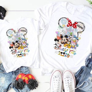 May include: White t-shirts featuring a colorful Disney design with Mickey Mouse, Minnie Mouse, Donald Duck, and Goofy. The design includes a castle, fireworks, and the text "Disney Trip 2020". Accessories include denim shorts, shoes, and plush toys.