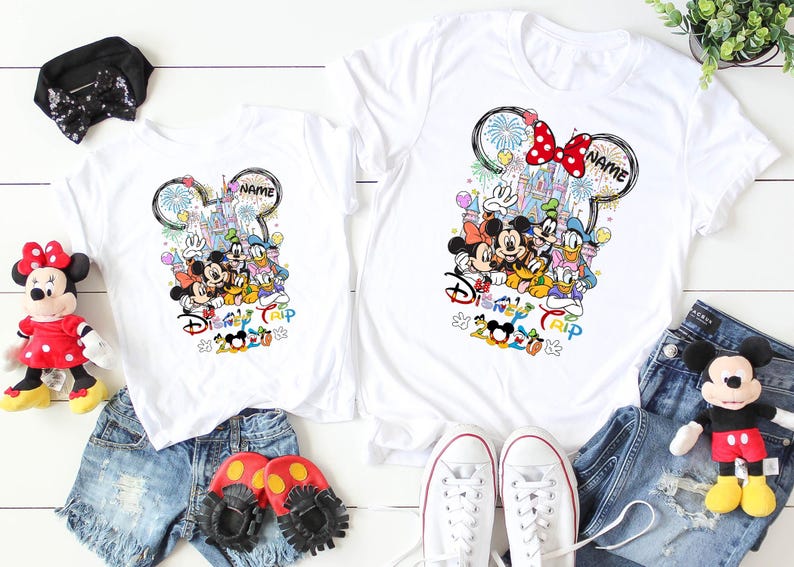 May include: White t-shirts featuring a colorful Disney design with Mickey Mouse, Minnie Mouse, Donald Duck, and Goofy. The design includes a castle, fireworks, and the text "Disney Trip 2020". Accessories include denim shorts, shoes, and plush toys.