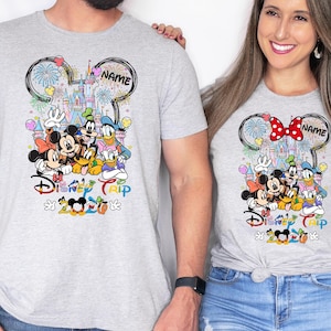 May include: Two light gray t-shirts featuring a Disney-themed design. The design includes Mickey and Minnie Mouse, Donald Duck, Goofy, and other characters, with a castle and fireworks. The text "Disney Trip" and "2023" are also visible.
