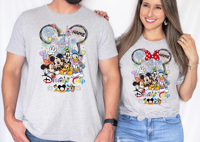 May include: Two light gray t-shirts featuring a Disney-themed design. The design includes Mickey and Minnie Mouse, Donald Duck, Goofy, and other characters, with a castle and fireworks. The text "Disney Trip" and "2023" are also visible.