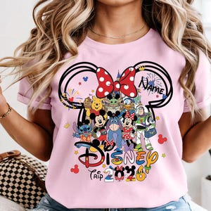 Peut inclure: T-shirt rose clair avec un motif sur le th&egrave;me de Disney. Le motif comprend des oreilles de Mickey Mouse avec un n&oelig;ud &agrave; pois rouges, divers personnages Disney et les mots "Disney Trip 2023". Le mot "Name" est &eacute;galement inclus.