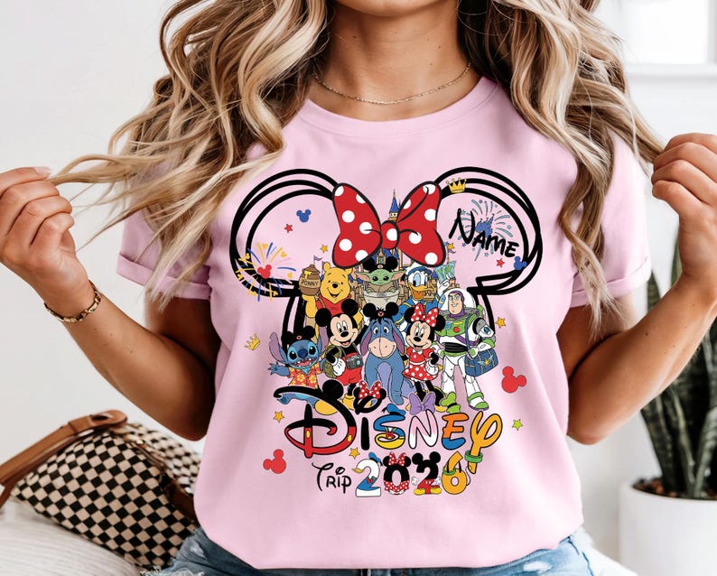 Peut inclure: T-shirt rose clair avec un motif sur le th&egrave;me de Disney. Le motif comprend des oreilles de Mickey Mouse avec un n&oelig;ud &agrave; pois rouges, divers personnages Disney et les mots "Disney Trip 2023". Le mot "Name" est &eacute;galement inclus.