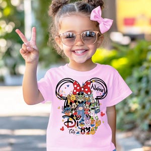 Peut inclure: Un t-shirt rose clair avec un motif Disney. Le motif comprend des oreilles de Mickey Mouse, divers personnages Disney et le texte "Disney Trip 2020". Le t-shirt est port&eacute; par un jeune enfant.