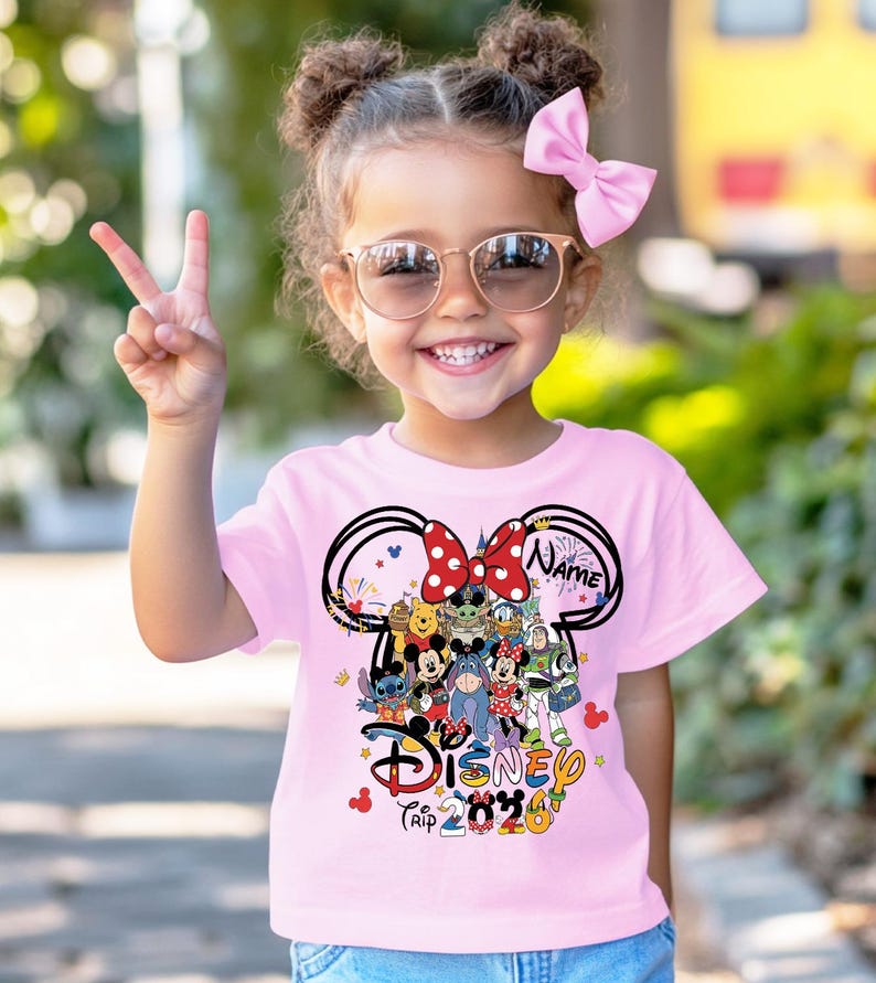 Peut inclure: Un t-shirt rose clair avec un motif Disney. Le motif comprend des oreilles de Mickey Mouse, divers personnages Disney et le texte "Disney Trip 2020". Le t-shirt est port&eacute; par un jeune enfant.