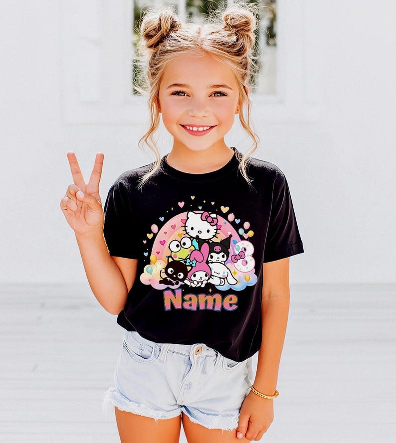 May include: A black t-shirt featuring a colorful graphic of Hello Kitty and friends, with a rainbow and hearts design. The word "Name" is printed in a pink and purple font. The shirt is worn by a young person.